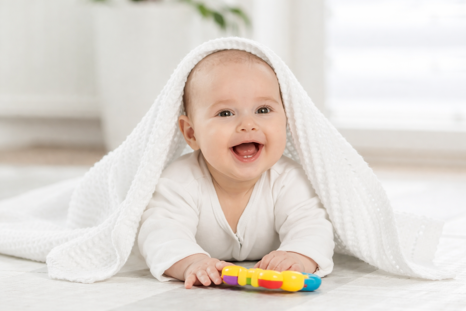 Lachendes Baby liegt auf dem Bauch unter einer weißen Decke und hält ein buntes Spielzeug.