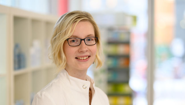 Mitarbeiterin einer Apotheke steht im Verkaufsraum vor Regalen mit Produkten.