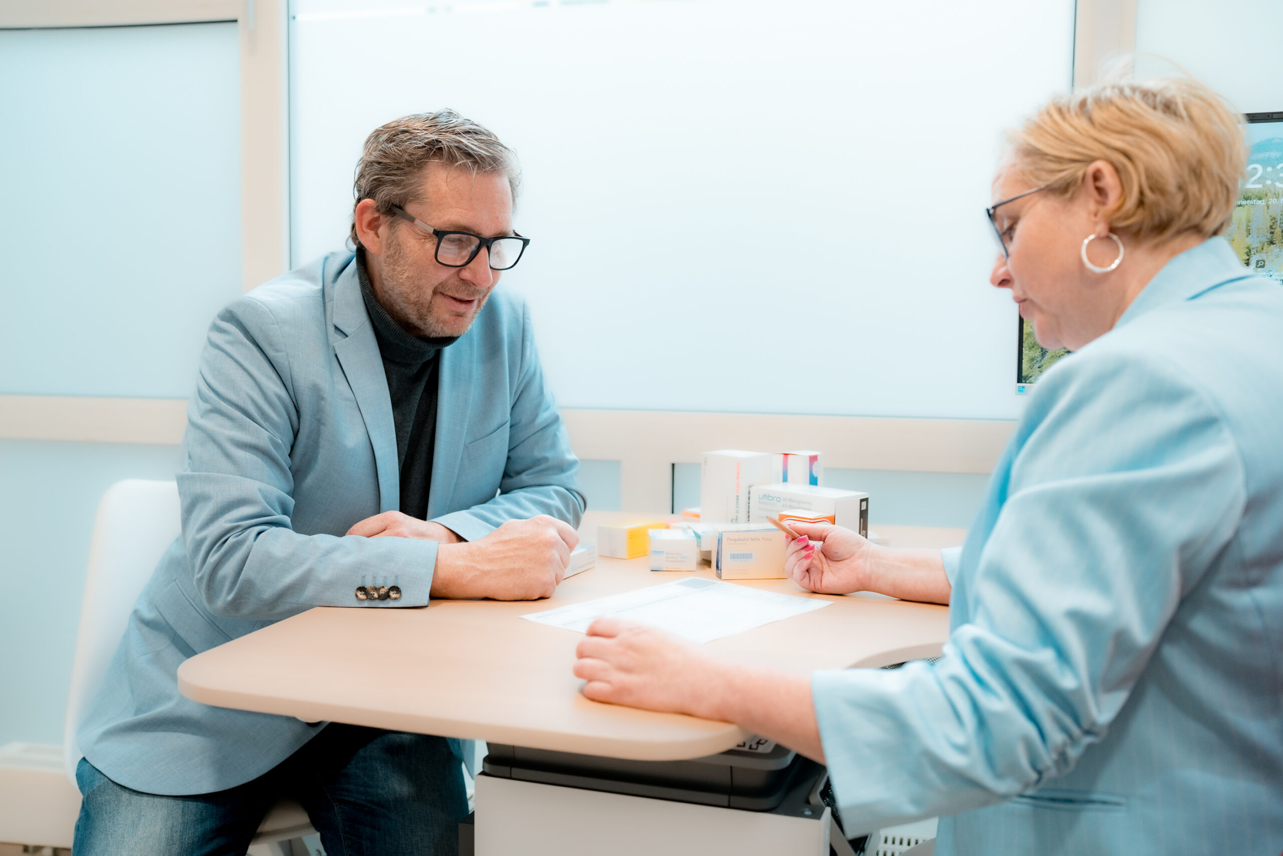 Mitarbeiterin einer Apotheke bespricht mit einem Kunden Medikamente und Unterlagen an einem Beratungstisch.